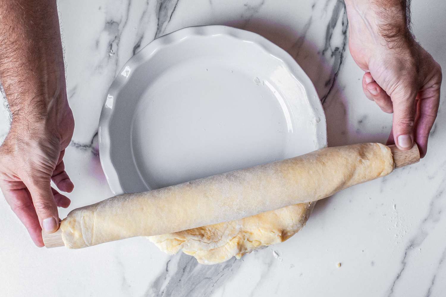 Hands rolling out dough into a pie dish using a rolling pin