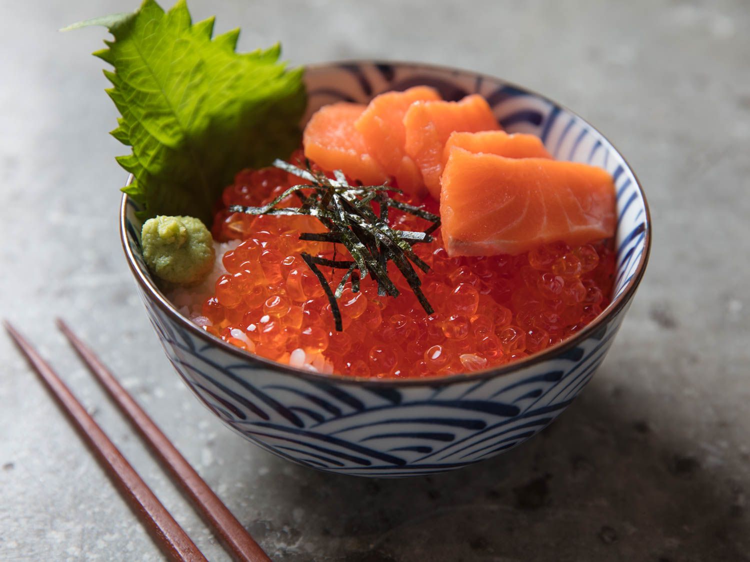 Close up of a bowl of ikura don, garnished with nori, wasabi, shiso leaf, and salmon sashimi. 