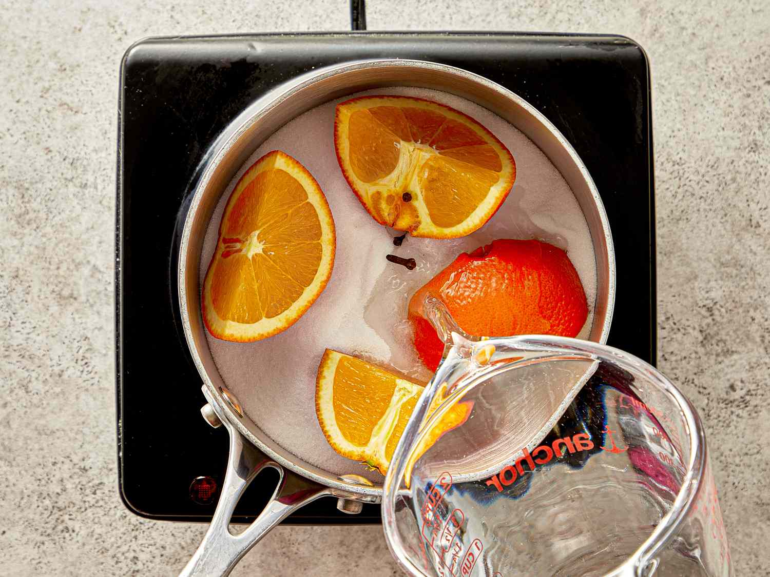 Orange slices and a peel in a pan of water on a stove with a measuring cup being used to pour additional water