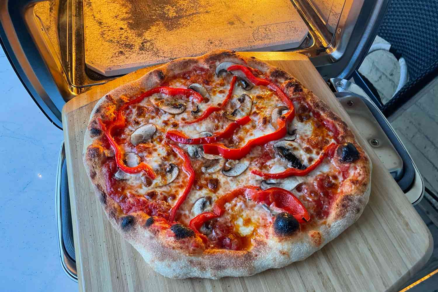 A freshly made pizza with mushrooms and red bell peppers on a wooden cutting board next to an oven