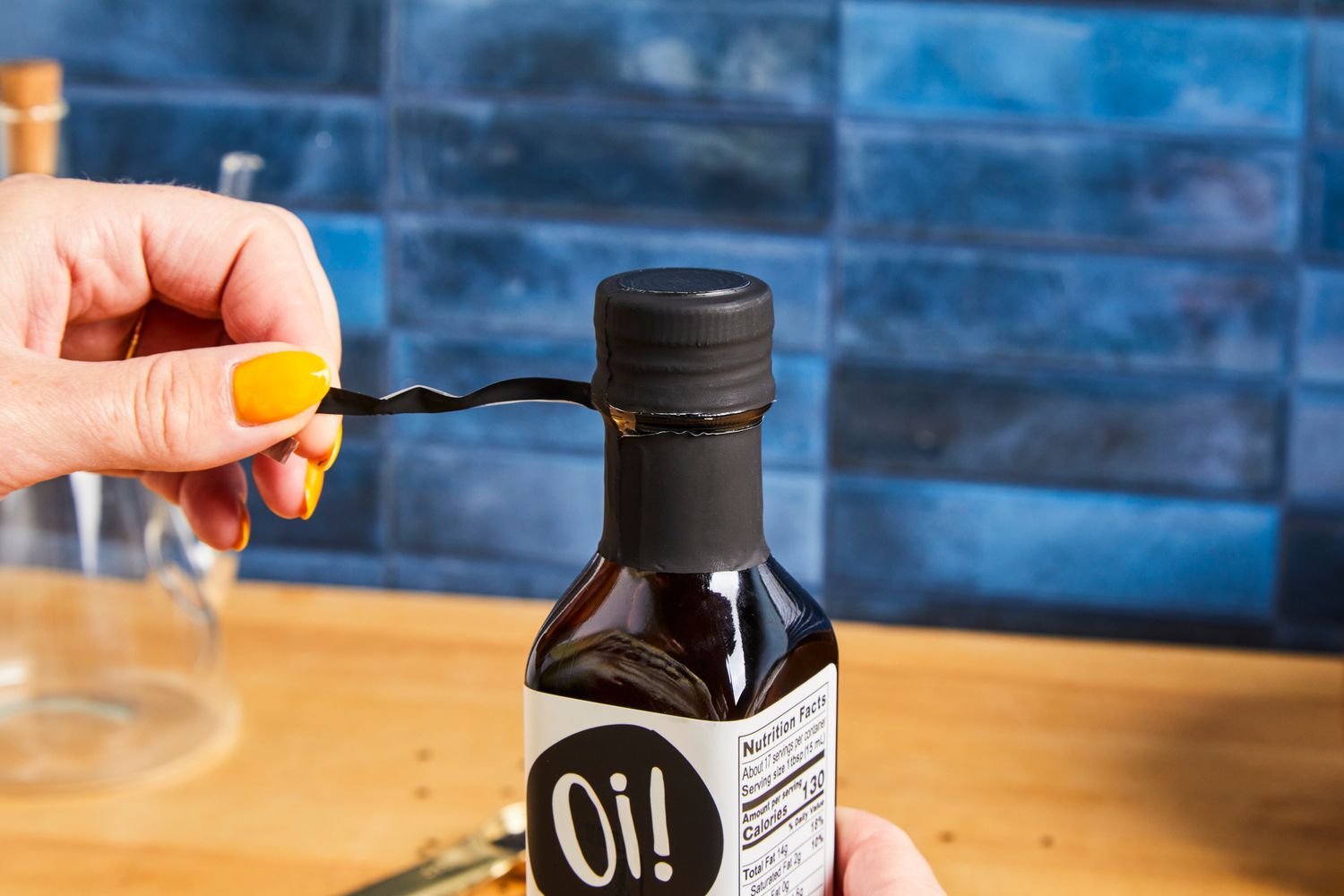 A hand peeling a strip from the cap of a bottle with "Oi!" branding on the label, background of blue tiles and wooden surface