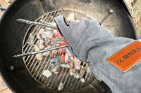 A person holds tongs above hot coals while wearing the Breeo Fire Pit Gloves