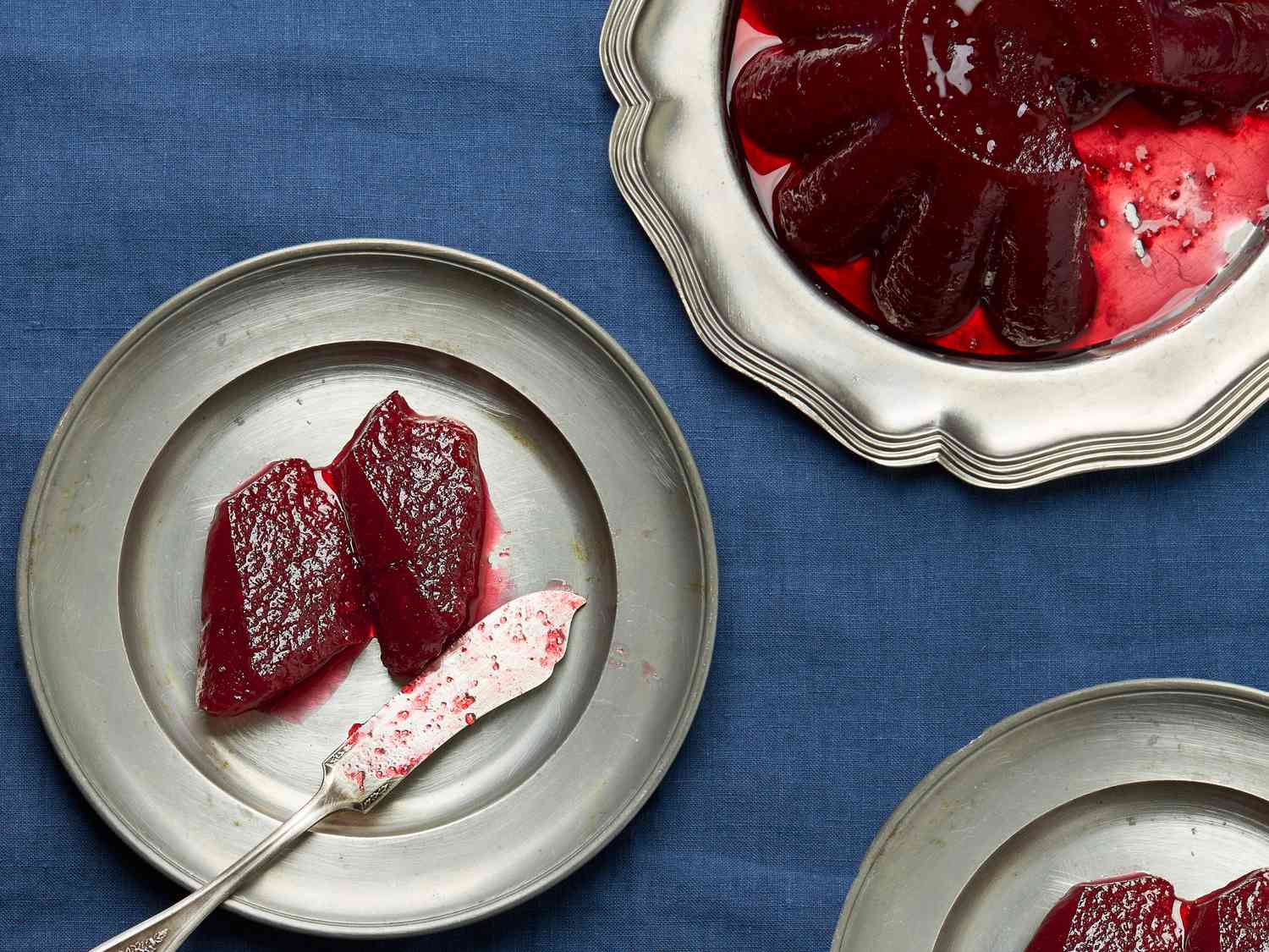 Small silver plates of serving of jellied cranberry sauce, with large platter of cranberry sauce in the top right. Platters sit on a blue fabric. 