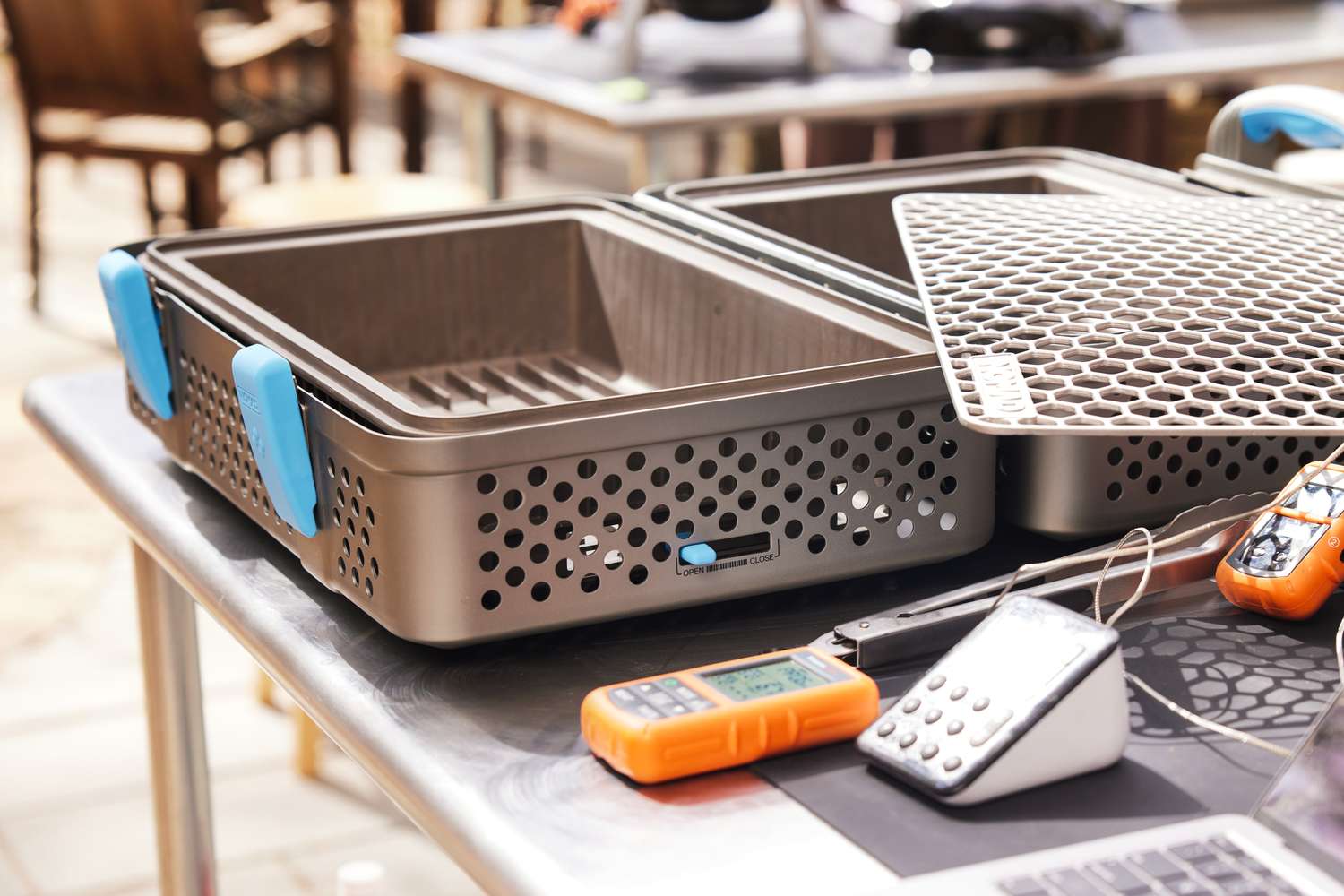 Kitchen equipment placed on a steel table including two metal perforated trays electronic devices and utensils