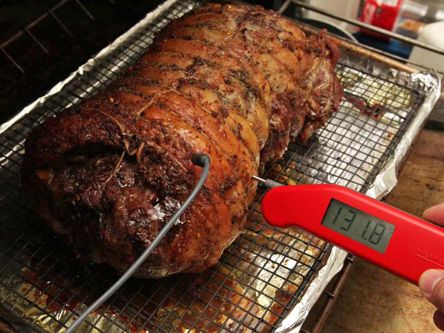A boneless, tied leg of lamb set on a wire rack in a foil-lined rimmed baking sheet. An instant-read thermometer registers 131.8 degrees Fahrenheit.