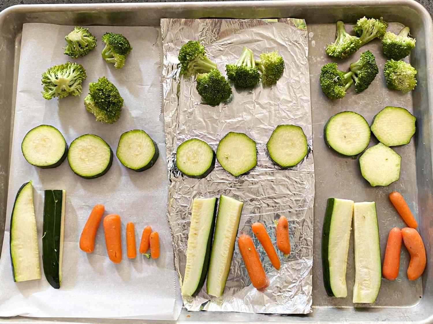 A baking sheet with rows of broccoli florets zucchini slices and carrot sticks placed on aluminum foil and parchment paper in sections
