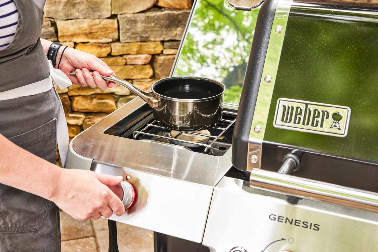 A person cooks food in a small pot on the Weber Genesis E-335 Gas Grill cooktop