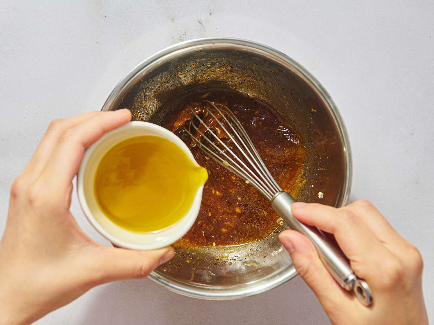 Hands pouring olive oil into a bowl with a whisk and a liquid mixture