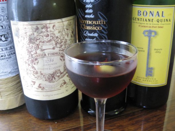 A vermouth cocktail in a cocktail glass with an olive. There are bottles of vermouth in the background.