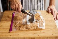 Person using a meat tenderizer to crush crackers in a resealable plastic bag on a cutting board