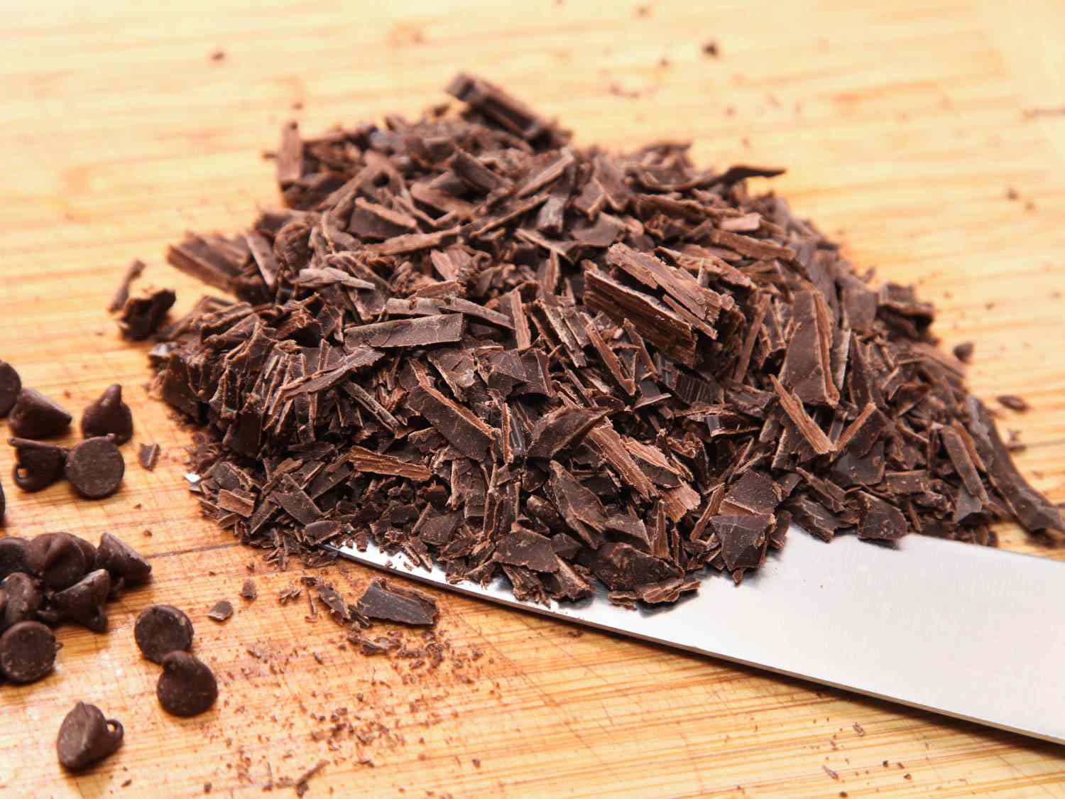 A pile of chopped chocolate on a cutting board next to a few chocolate chips.