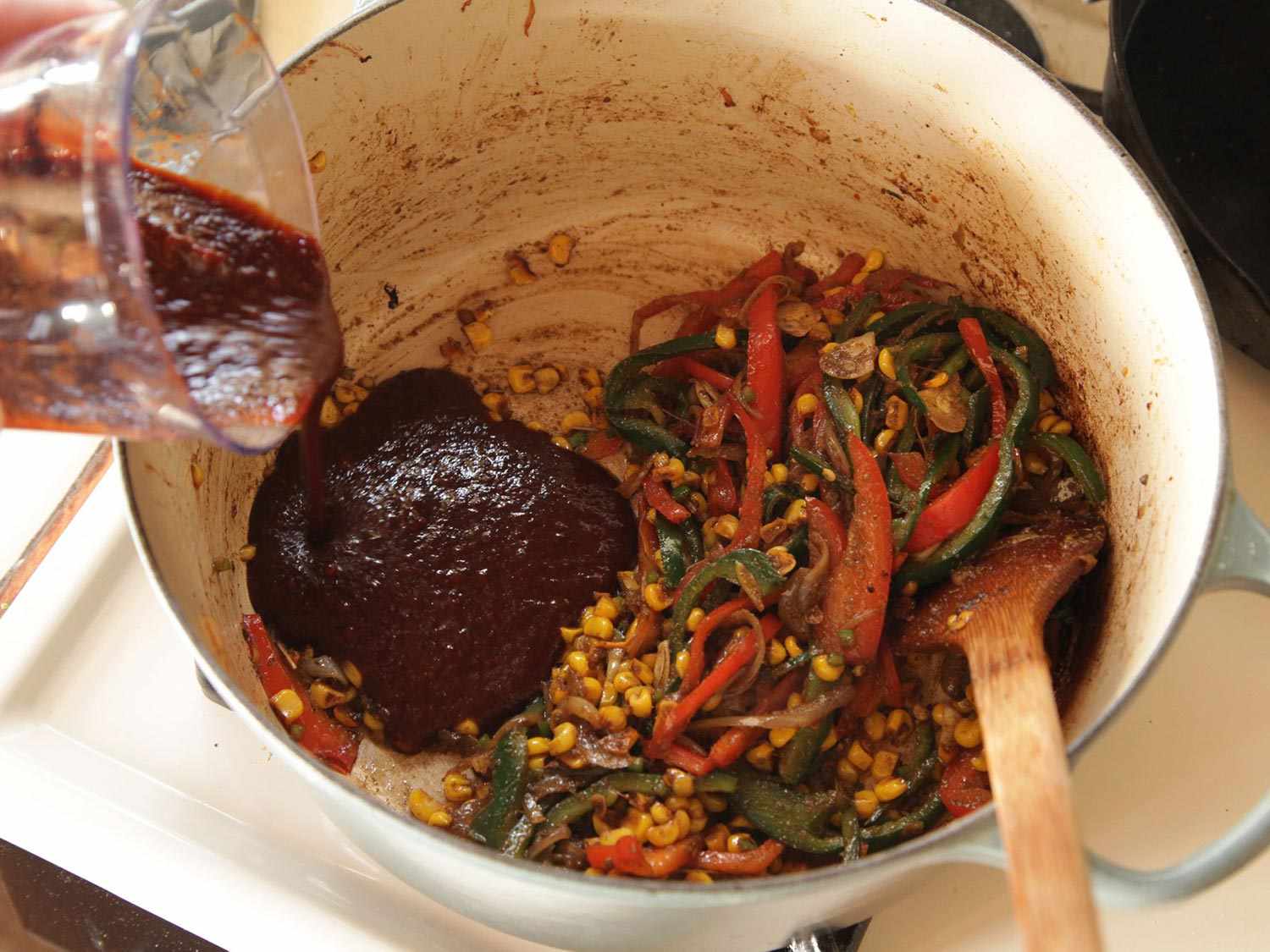 Pouring dark red chile sauce into Dutch oven with sauteed vegetables