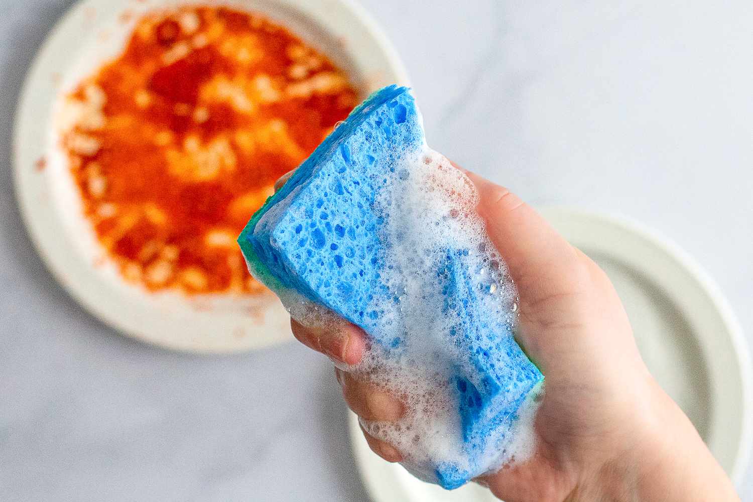 a person using a soapy sponge to clean a dish with pasta sauce on it. 