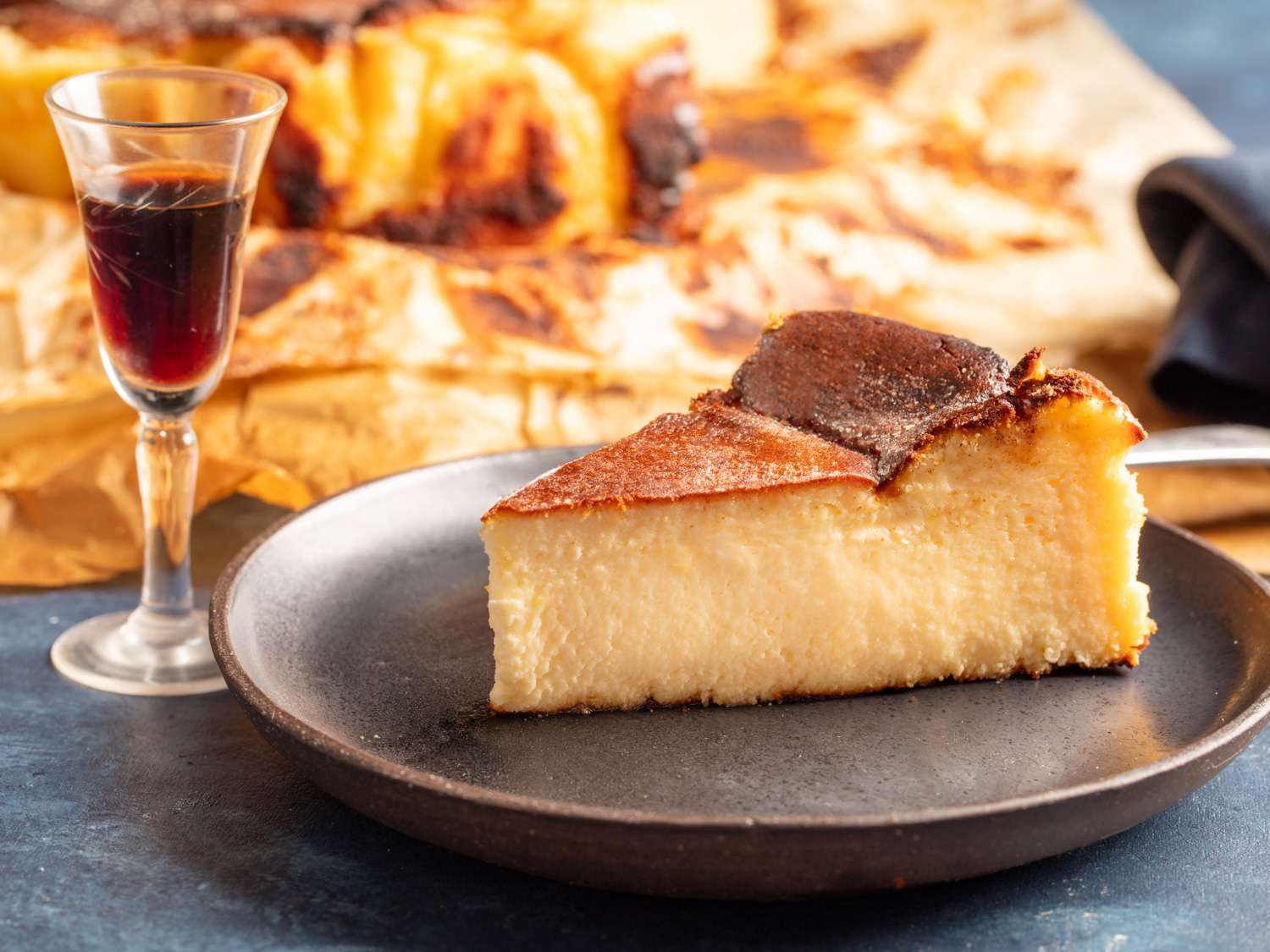 A slice of Basque cheesecake resting on a plate with a glass of port off to the side.