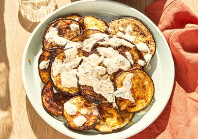 Grilled eggplant slices topped with a creamy white sauce served in a bowl on a wooden table beside a red cloth napkin
