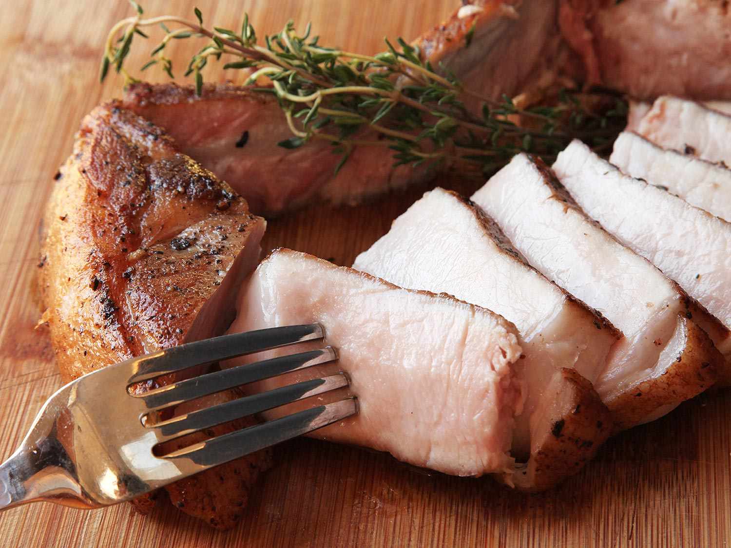 A bone-in cooked pork chop on a cutting board that's been sliced. A fork is in one slice. There is a sprig of fresh thyme next to the pork slices.