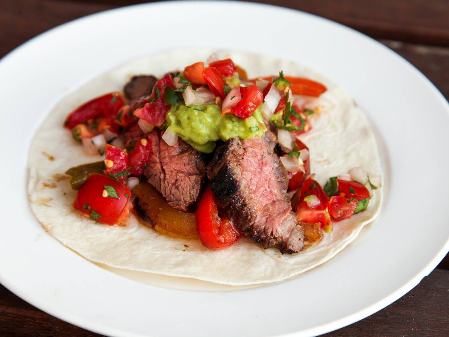 A white plate holding a flour tortilla topped with steak fajitas, grilled peppers and onions, guacamole, and pico de gallo.