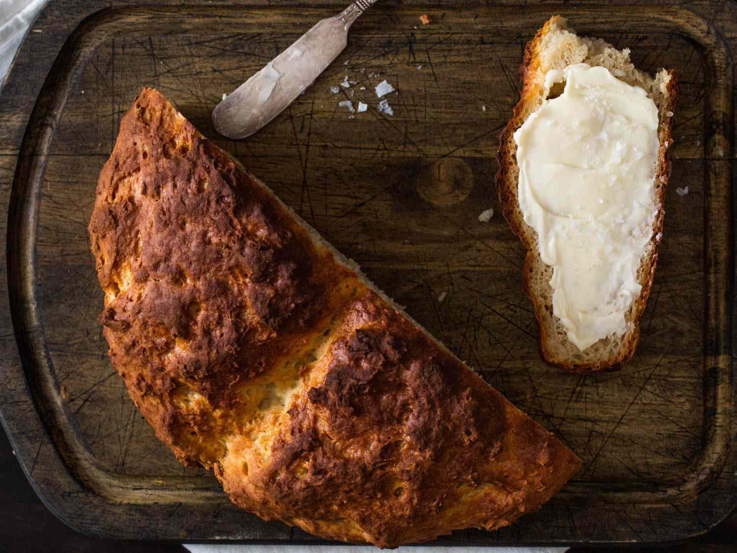 Overhead shot of half a loaf of Irish soda bread on a wooden board, next to a slice of bread spread with butter.