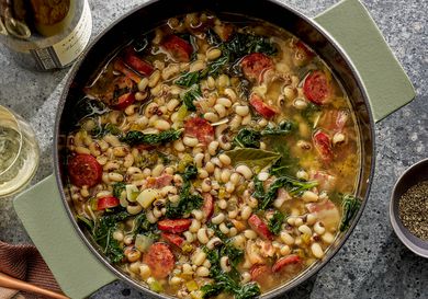 Pot of black eyed peas, kale and sausage stew, surrounded by a printed napkin, bottle and glass of wine on a stone surface 