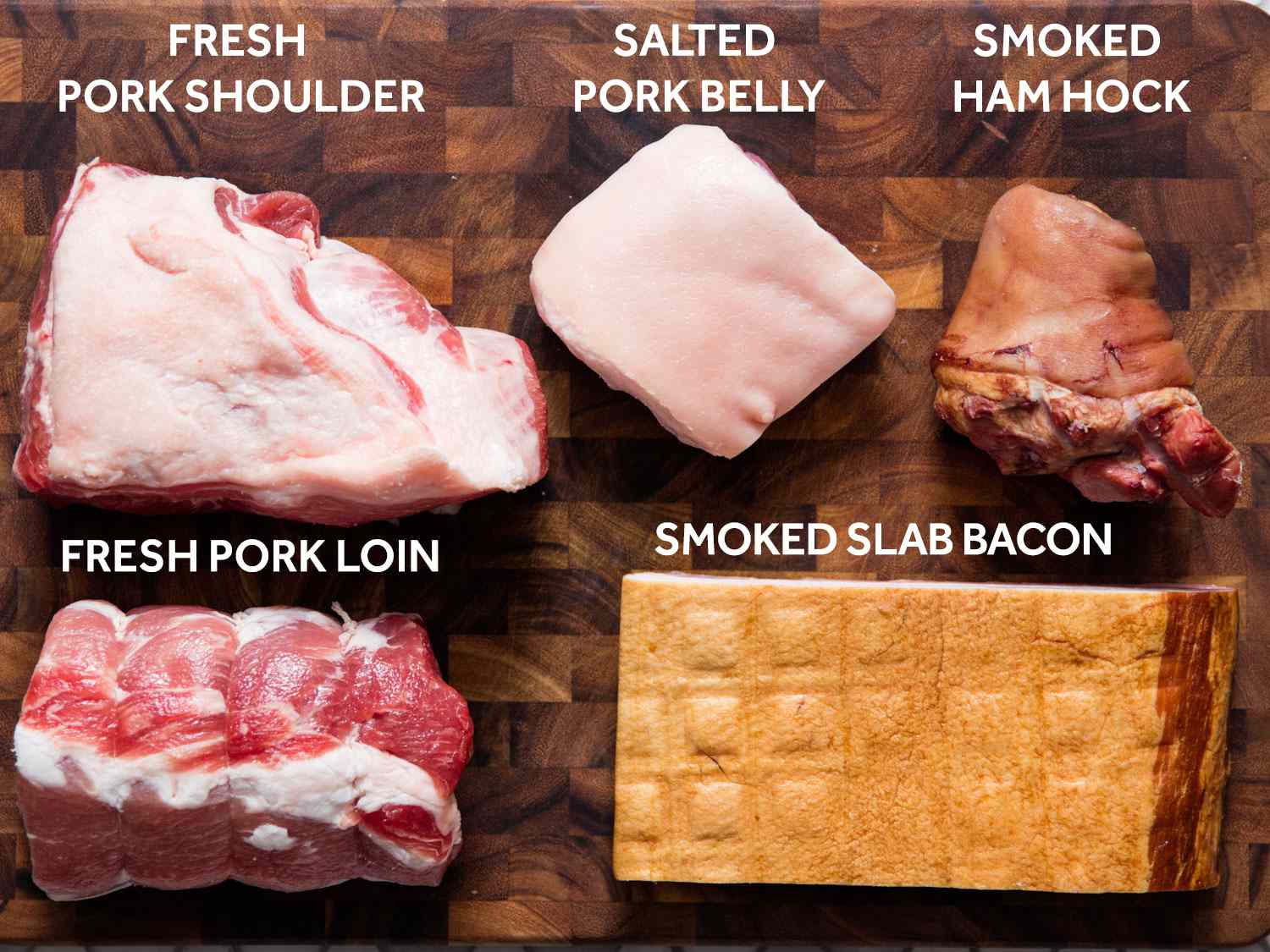 All of the meats (aside from sausage) needed for choucroute garnie, laid out on a cutting board and labeled: fresh pork shoulder, salted pork belly, smoked ham hock, fresh pork loin, smoked slab bacon.