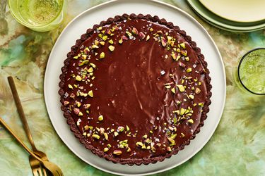 Whole Dubai Chocolate Tart topped with pistachio, on green tablecloth with 2 glasses of water, and stacks of plates in the corner 