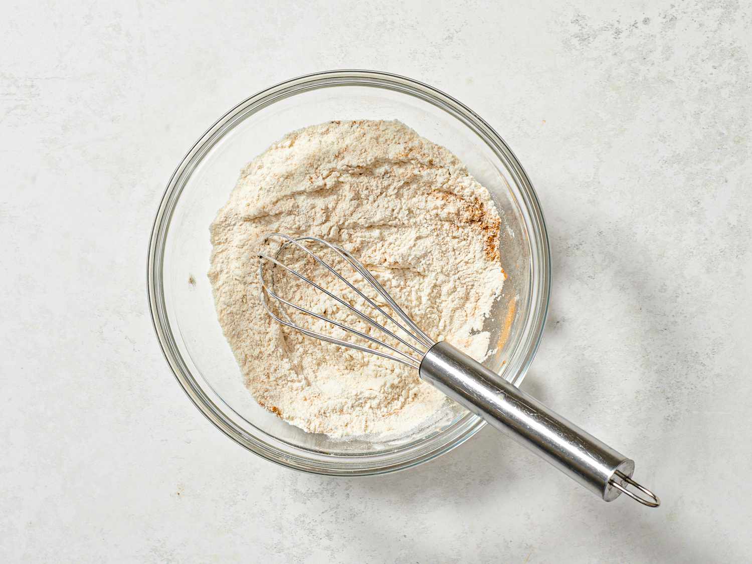 Flour, cinnamon, salt, and baking soda whisked together in a medium glass bowl. The whisk is still inside of the bowl.