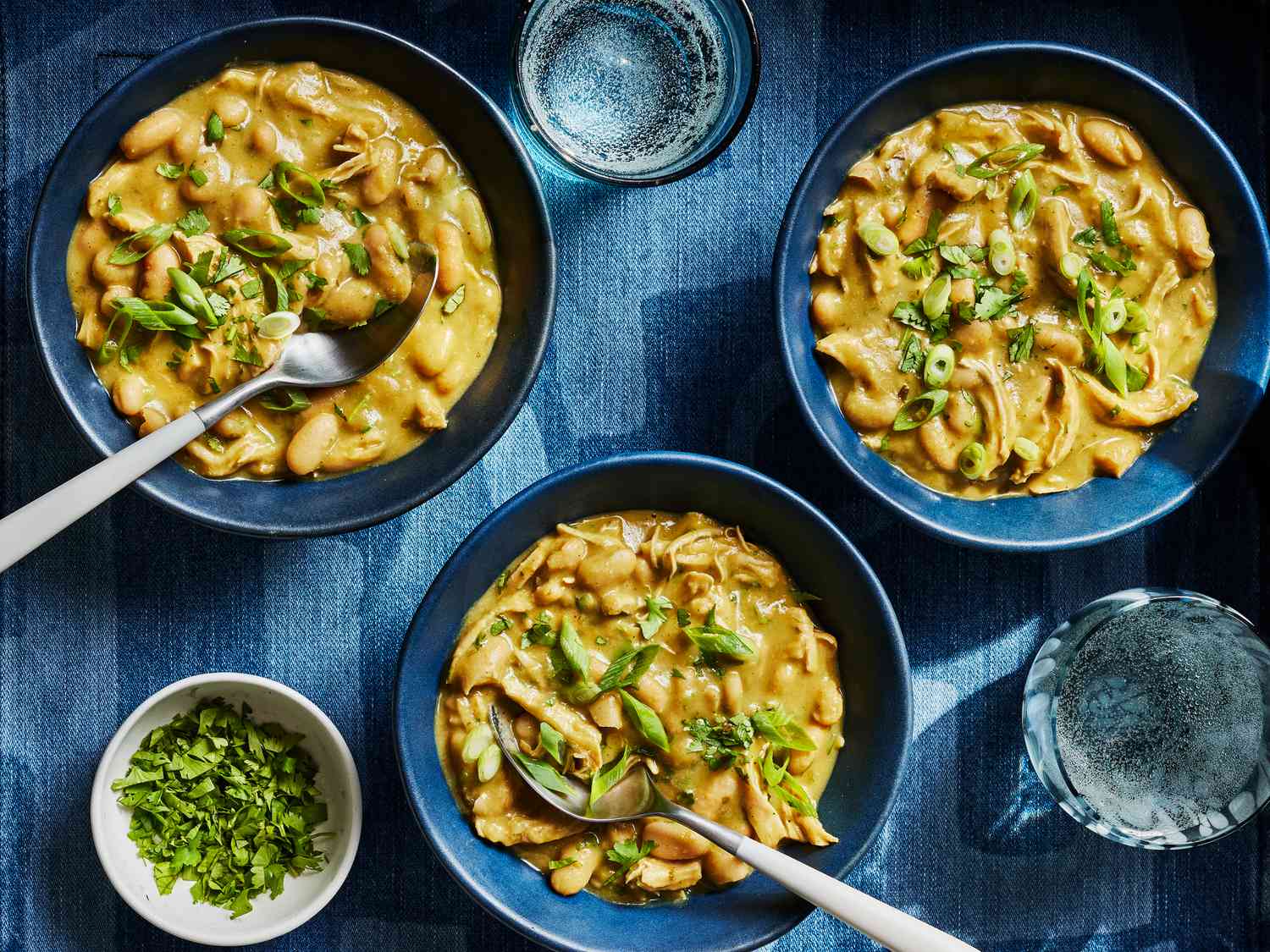 3 blue bowls of chicken, on a chambray textile with 2 glasses of sparking water 
