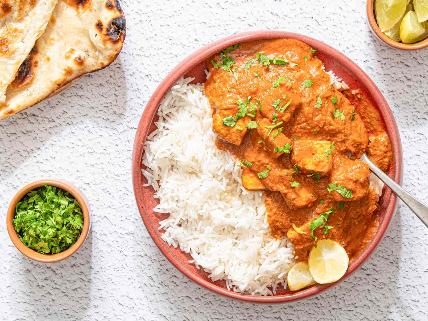 A bowl of tofu tikka masala with rice and garnished with fresh herbs accompanied by naan bread lime wedges and a small bowl of chopped herbs