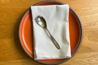 A spoon rests on a folded napkin placed atop a round plate on a wooden surface