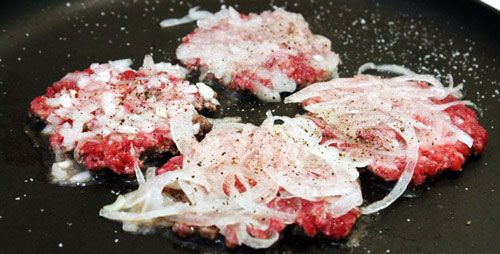 slider patties in a skillet, topped with thinly sliced onion.