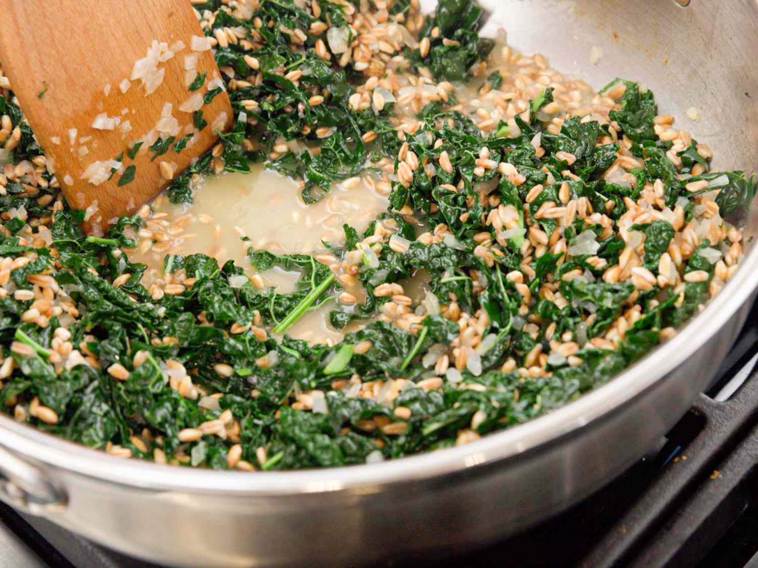 Kale sauteeing into onion and farro in skillet being mixed with wooden spoon 