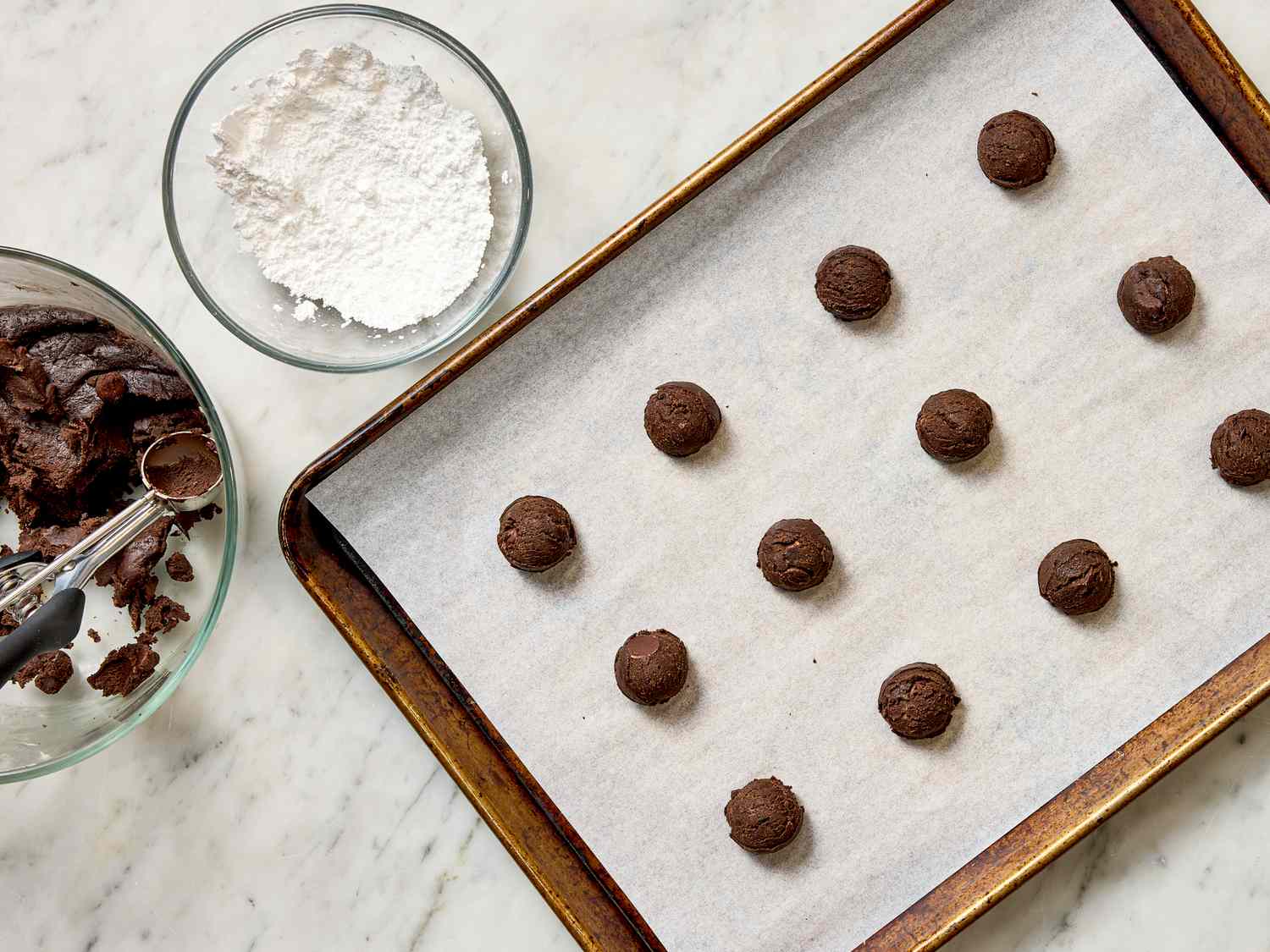Baking tray with chocolate cookie dough balls adjacent bowls of dough and powdered sugar