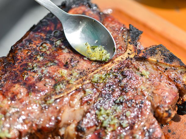 Close-up of melted herb butter being spooned onto the surface of a charred porterhouse steak. 