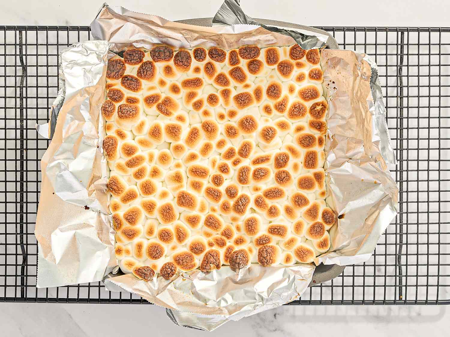 A square smores brownie topped with toasted marshmallows on a wire rack and aluminum foil