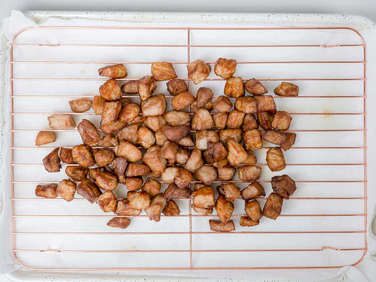 Fried pork shoulder pieces sitting on a copper rack set above layers of paper towel.