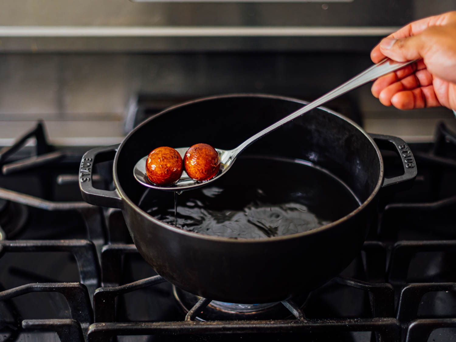 Using a slotted spoon to lift two dark brown gulab jamun from a pot of hot oil.