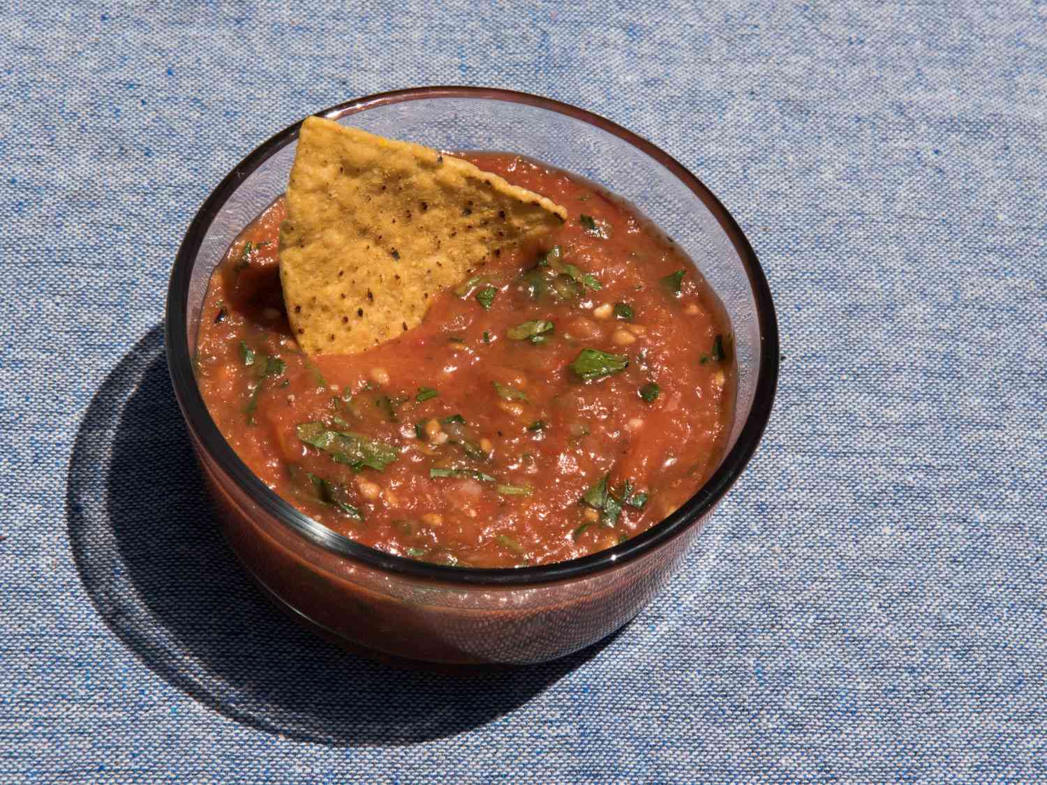 A glass bowl with roasted tomato salsa with a chip in it.