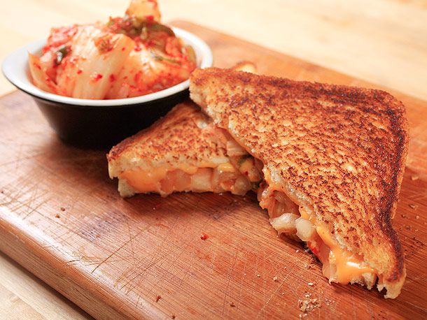 A grilled kimchi and cheese sandwich split in half next to a bowl of kimchi.