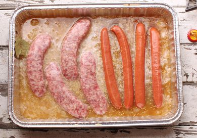 Overhead view of sausages and hot dogs partially submerged in a disposable aluminum pan filled with a flavorful braising liquid.