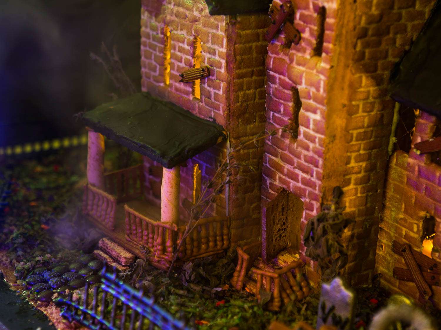 Angled view of front of haunted gingerbread house, with spooky dim lighting, with falling-down railings, gingerbread tombstones, and green-dyed coconut lawn visible