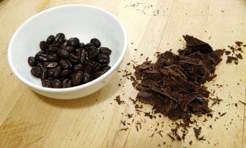 Bowl of coffee beans next to chopped chocolate on a wooden cutting board.