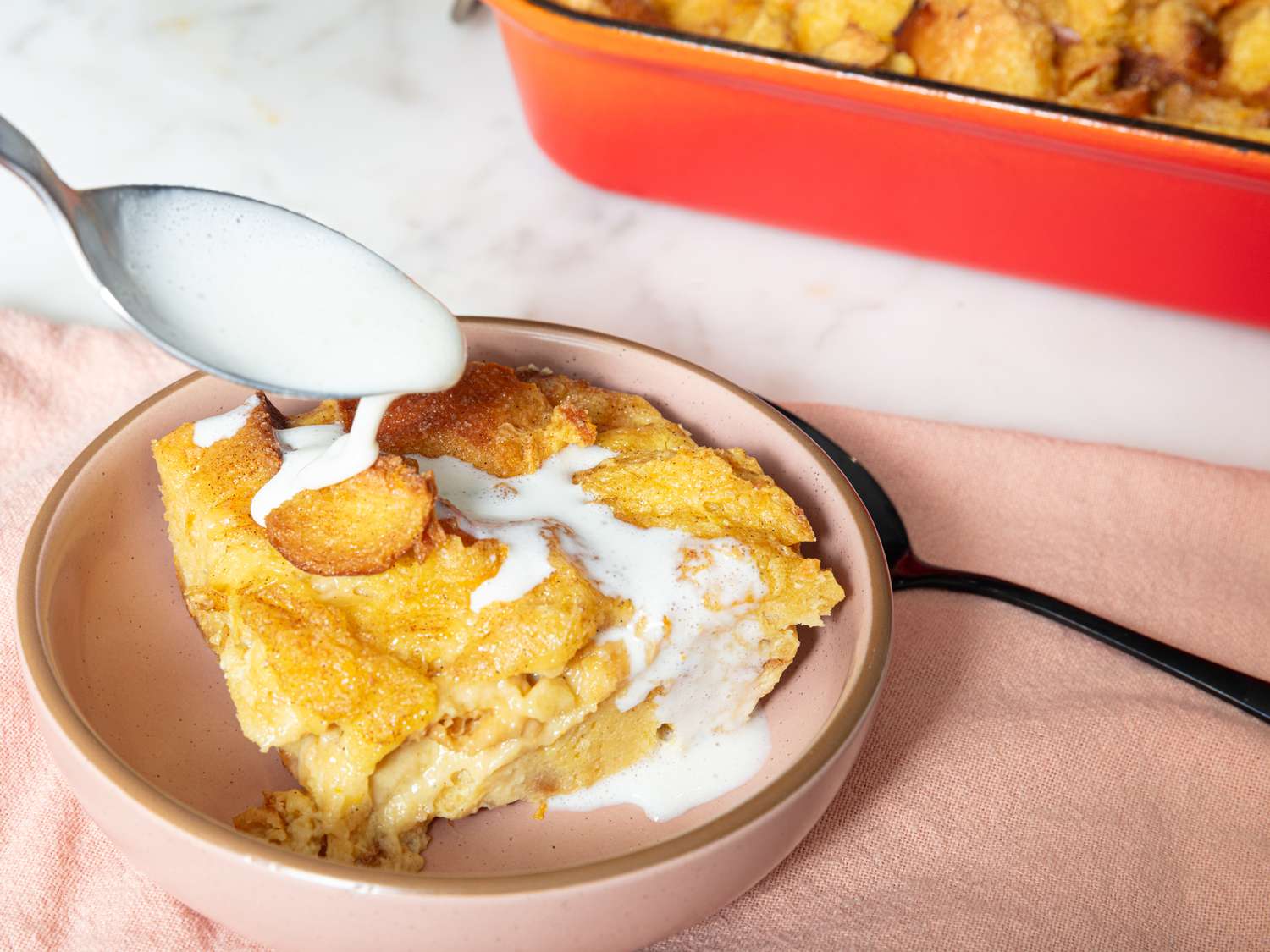 Piece of bread pudding in a peach bowl, with a spoon drizzling creme Anglaise on top. Bowl is on a peach textile, on a marble surface, and a red tray of bread pudding in the background. 