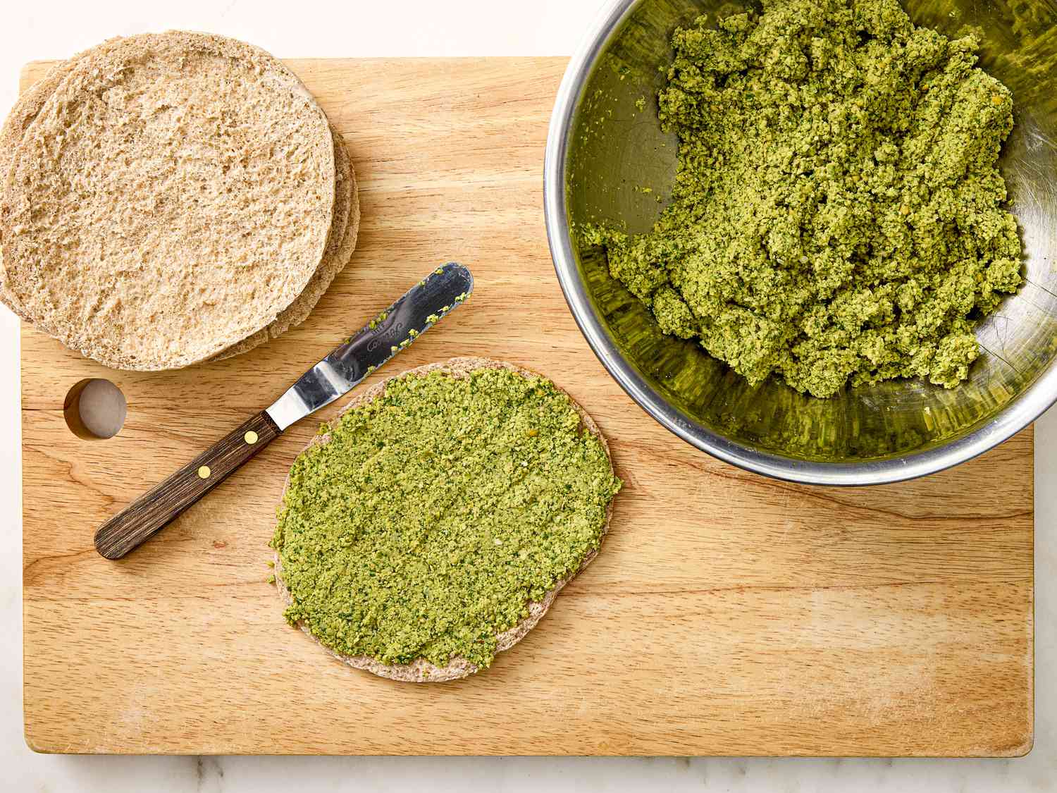 Falafel spread across a pita on a cutting board, next to a stack of pitas and a bowl of falafel mix 
