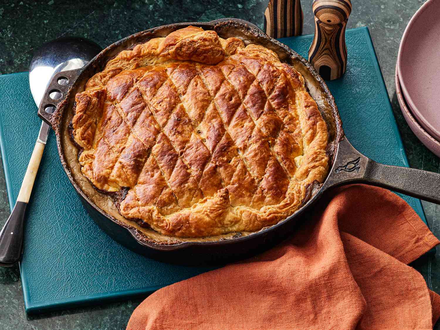 Mushroom Pot Pie in a skillet on a blue place mat, with a rust colored napkin, soft pink plates, and a serving spoon to the side 