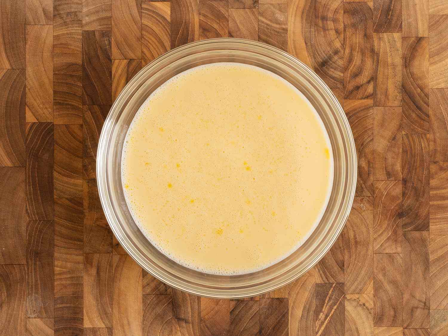 Eggs and milk whisked together in a glass bowl on a wood counter.