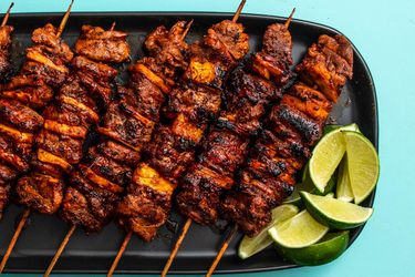 A black platter of charred al pastor skewers and a pile of lime wedges for last-minute drizzling.