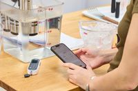 Person using a phone to control a Anova Culinary Precision Cooker 3.0 with a container of water and measuring tools on a nearby table