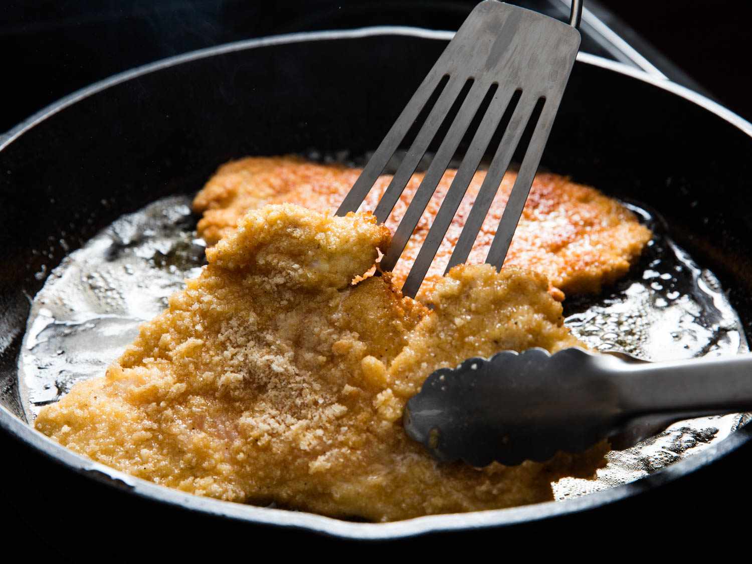 Flipping a chicken cutlet with the help of tongs and a slotted fish turner.