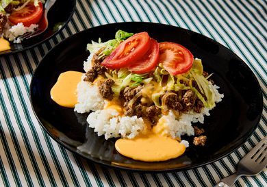 A plate of taco rice with beef, lettuce, and sliced tomatoes on top of white rice, served on a striped tablecloth