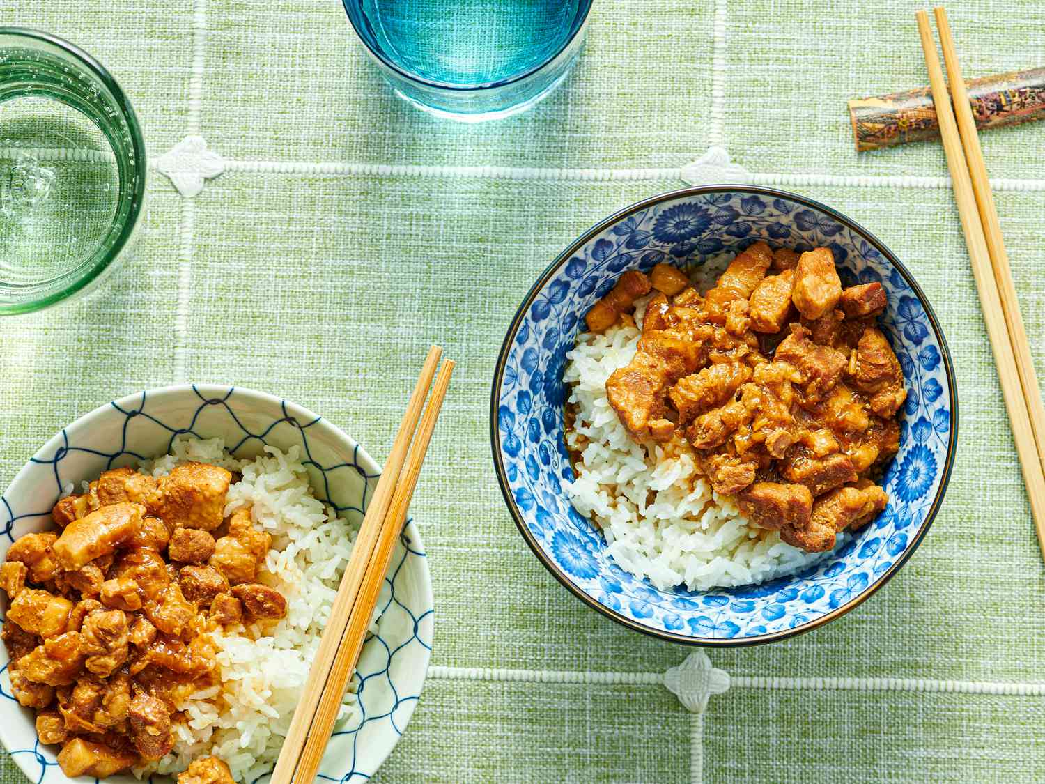 2 bowls of minced pork on white rice in a blue bowl and a white bowl, with chop sticks and glasses of water on a green tablecloth